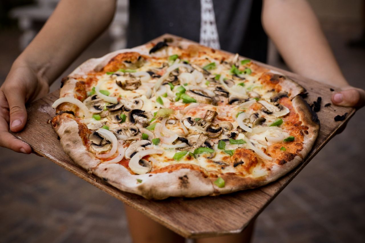Large pizza on a wooden tray with two arms holding it.